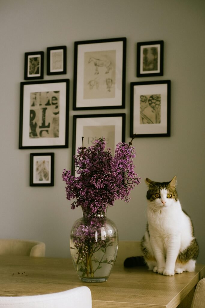 A cozy interior scene featuring a cat sitting next to a vase of lilacs with framed art on the wall.